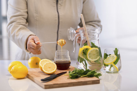 Female hand holding a dripping honey dipper over a honey jar on a wooden cutting table with lemons. Selective focus. Homemade remedies concept.の写真素材