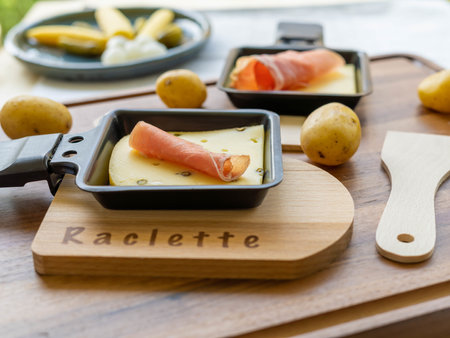 A selective focus shot of raclette pepper cheese with parma ham in a grill pan on a wooden boardの写真素材