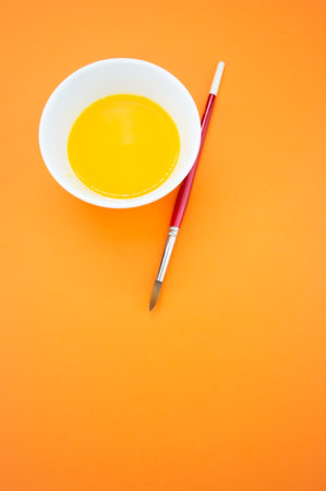 A vertical shot of a yellow watercolor in a bowl and a paintbrush isolated on an orange backgroundの写真素材