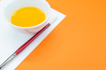 A closeup shot of a yellow watercolor in a bowl and a paintbrush on a sheet of paperの写真素材