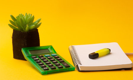 A selective focus shot of highlighter on a journal, calculator, and a plant on a yellow backgroundの写真素材