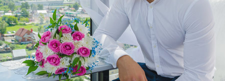 A young man in a white shirt, the groom, sits by the window with a table on which lies a bridal...