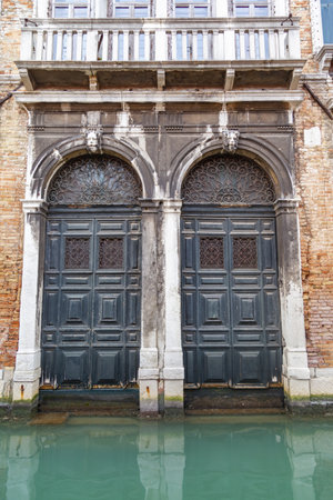 A vertical shot of an old building with wooden doors surrounded by a canal in Venice, Italyの写真素材