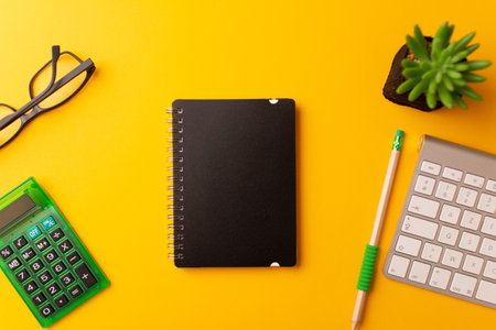 A top view of a journal, calculator, a pencil, keyboard, and a plant on a yellow backgroundの写真素材