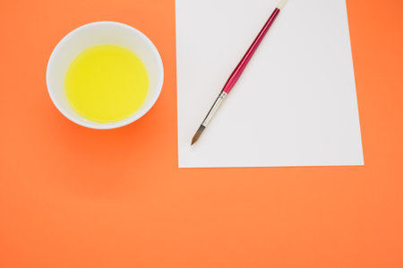 A high angle shot of yellow paint in a container with a brush and paper on an orange surfaceの写真素材
