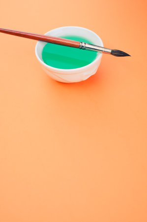 A vertical shot of a bowl of green paint and a paintbrush on an orange surfaceの写真素材