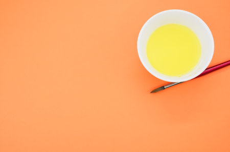 A top view of yellow paint in a container with a brush on an orange surface
