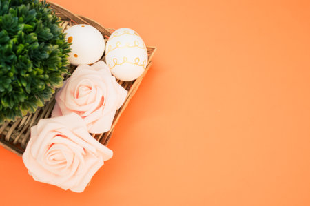 A basket with a small potted tree, roses, and easter eggs on an orange surfaceの写真素材