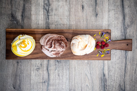 A top view shot of three meringue cookies with different flavors set on a narrow brown wooden cutting board on top of a light gray wooden surface.の写真素材