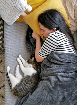 A young Caucasian woman sleeping on a couch with a cat under a warm blanket