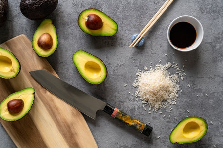 A top view of fresh avocados, a cutting board and knife, rice, and chopsticks on a grey surfaceの写真素材