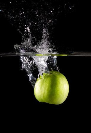 A vertical shot of a green apple in water with splashes on a dark isolated backgroundの写真素材