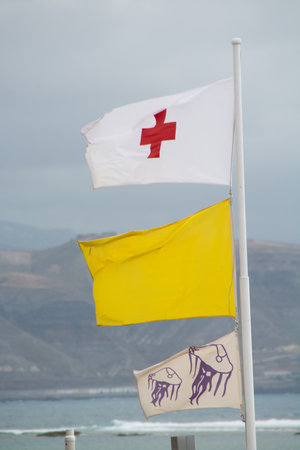 A vertical shot of the Red Cross flag, yellow flag, and a Jellyfish Warning flag waving in the windの写真素材