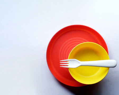 A top view of a red plate, a yellow bowl and a white fork on a white surface with copy spaceの写真素材