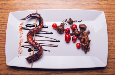 A top view of an octopus tentacle on a stick served with tomatoes and mushrooms on a white plate