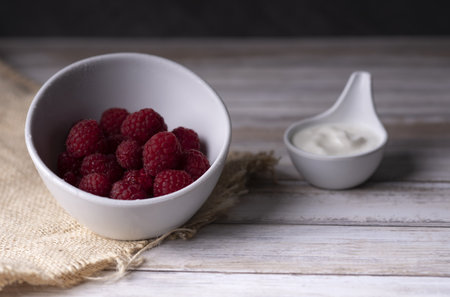 A bowl of raspberries served with natural white cream on a wooden tableの写真素材