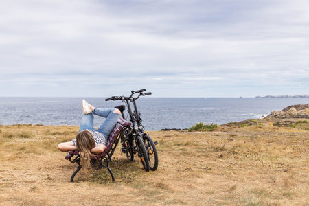 Caucasian blonde woman lying on a wooden bench relaxing in front of the sea. Relaxing girl after biking on vacation in the coast.の写真素材