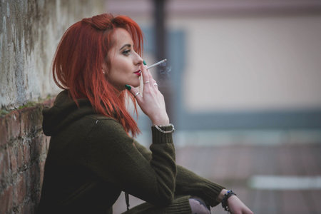 A red-haired young female in a beautiful winter dress smoking in front of a brick wallの写真素材