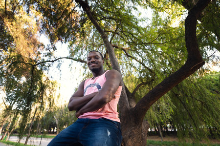 A low angle shot of a young handsome Ðexican male leaning on a tree in a parkの写真素材