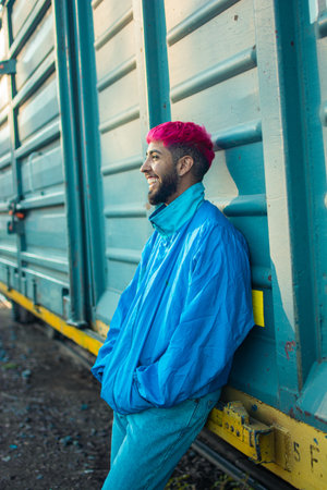 A handsome young man with stylish pink-dyed hair wearing a retro jacket leaning against a railway...