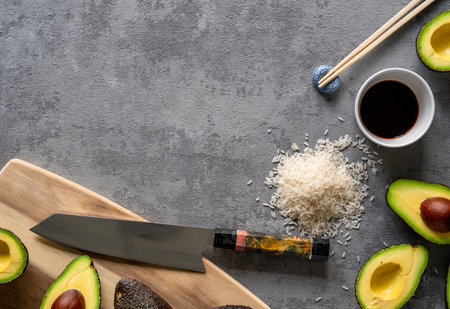 A top view of fresh avocados, a cutting board and knife, rice, and chopsticks on a grey surface,...