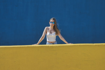 A young attractive girl from Spain wearing sunglasses behind a yellow wall with a blue background