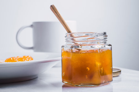 A closeup of an open jar of golden honey with honey dipper in it and ceramic teacup in the...