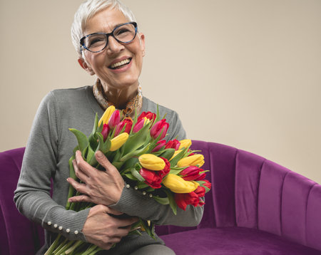 A beautiful senior female in eyeglasses holding tulips while sitting on a purple sofaの写真素材