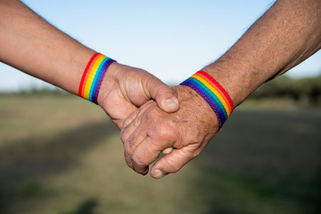 A closeup shot of a gay couple holding each other's hands with LGBT bracelets on a blurred...