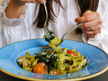 Close-up image of a woman eating green pasta with shrimps, cherry tomatoes and arugula. Ready to eat food served on blue ceramic plate.の写真素材