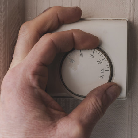 mans hand turning up the central heating temperature on a thermostat fixed to the the wall of his...
