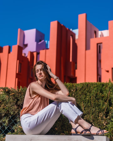A young attractive woman from Spain in a salmon-colored shirt and white pants sitting on a patio in front of geometric red buildingsの写真素材