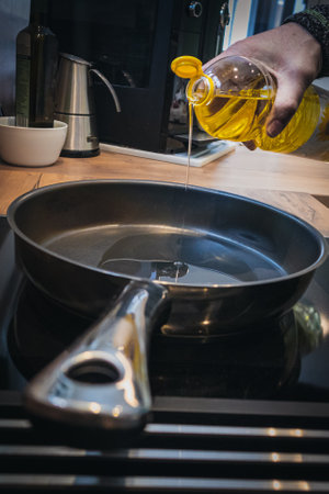 A pan on a stove and a bottle of oil in the hand of a cook, pouring oil in the pan. Demonstrating the start of the cooking in a kitchen.の写真素材