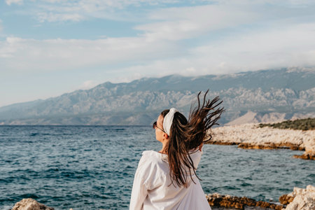 A young Caucasian woman in white long sleeve and sunglasses flipping her hair near the seaの写真素材