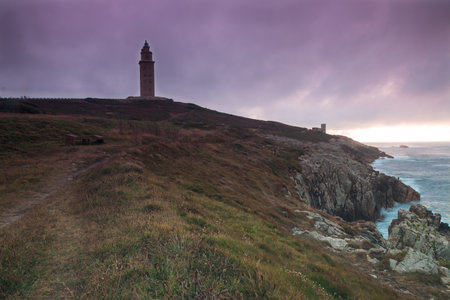 A mesmerizing view of the sea with a lighthouse on the hill under a beautiful sunset skyの写真素材