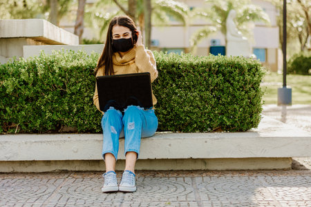 A young Argentinian female student wearing a yellow sweater sitting on a bench with laptop on her legsの写真素材