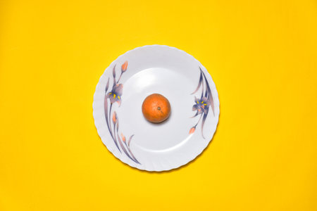 An orange on a white plate with floral patterns isolated on a yellow backgroundの写真素材