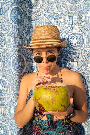 An attractive female in a swimsuit, sunglasses and hat drinking a fresh coconut cocktail