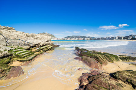 A beautiful view of the wavy ocean hitting the rocky La Concha beach under the blue skyの写真素材
