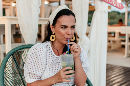 Joyful young woman holding drinking glass with refreshing cocktail and drinking straw. Attractive young woman in bar in summer.の写真素材