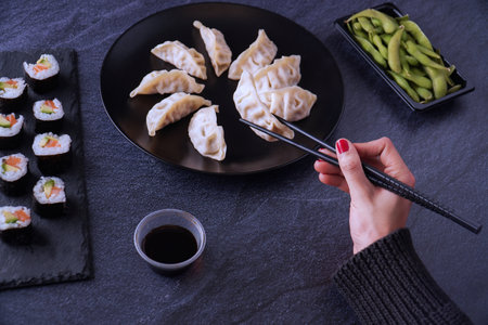 A high angle shot of a female hand about to take chicken gyozas, the salmon and avocado makis, endamame beans, and soy sauce served on the tableの写真素材