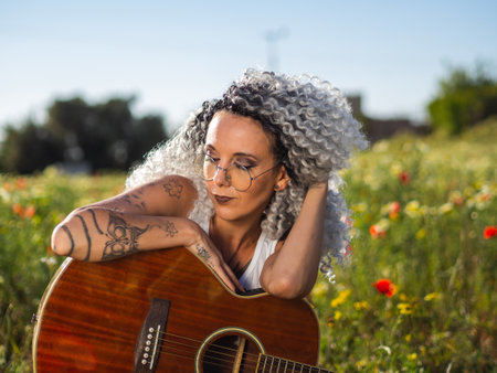 A cute Spanish woman with white curly hair and cool tattoos sitting in a meadow with a guitarの写真素材
