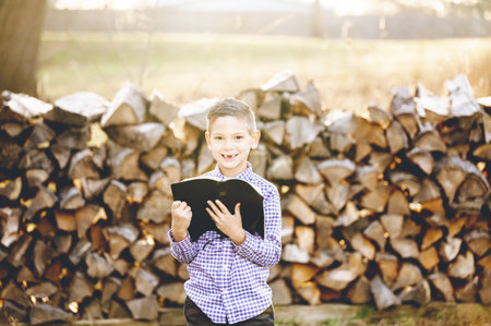 A little boy reading the holy bible while standing in front of stacked firewood in a fieldの写真素材