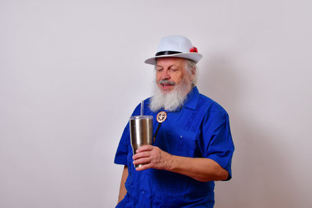 A happy old man with white fedora hat, blue guayabera shirt, and bolo tie holding insulated drink...