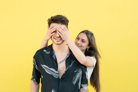 A beautiful shot of a girl covering her boyfriends' eyes isolated on a yellow backgroundの写真素材