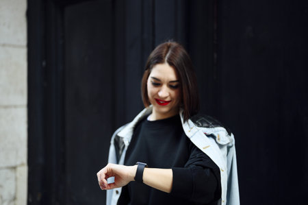 A cheerful attractive Spanish female looking at her wristwatch and posing on the black wall backgroundの写真素材