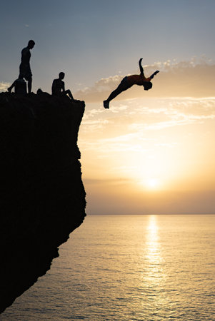 A silhouette of a man jumping off a cliff into the ocean at sunsetの写真素材