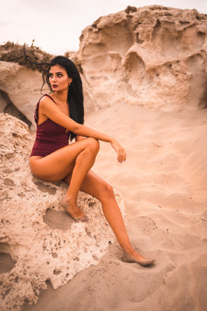 A young brunette Caucasian woman in a maroon swimsuit sitting on the beach of Cabo de Gata and posingの写真素材