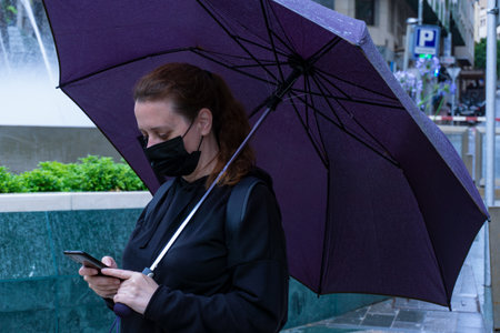 A caucasian female in a black protective mask with a purple umbrella scrolling on her phoneの写真素材