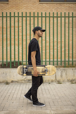 A young caucasian male from Italy in a black shirt and cap holding a skateboard in his hand on the streetの写真素材